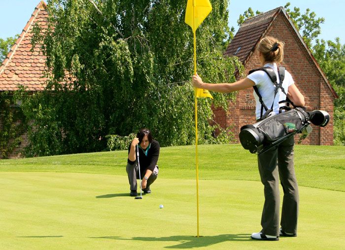 Zwei Frauen spielen Golf auf einem grünen Golfplatz, eine hält die Fahne, die andere zielt auf das Loch.