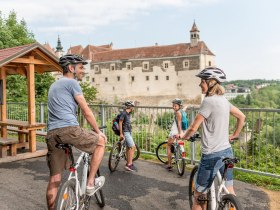 Eine Familie genie&szlig;t einen sonnigen Tag beim Radfahren in der malerischen Umgebung. Die sanften H&uuml;gel und die beeindruckende Burg im Hintergrund schaffen eine perfekte Kulisse f&uuml;r unvergessliche Erlebnisse in der Natur.