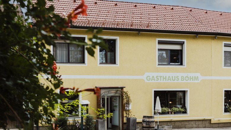 Gelbes Gasthaus mit roten Dachziegeln und Pflanzen vor dem Eingang.