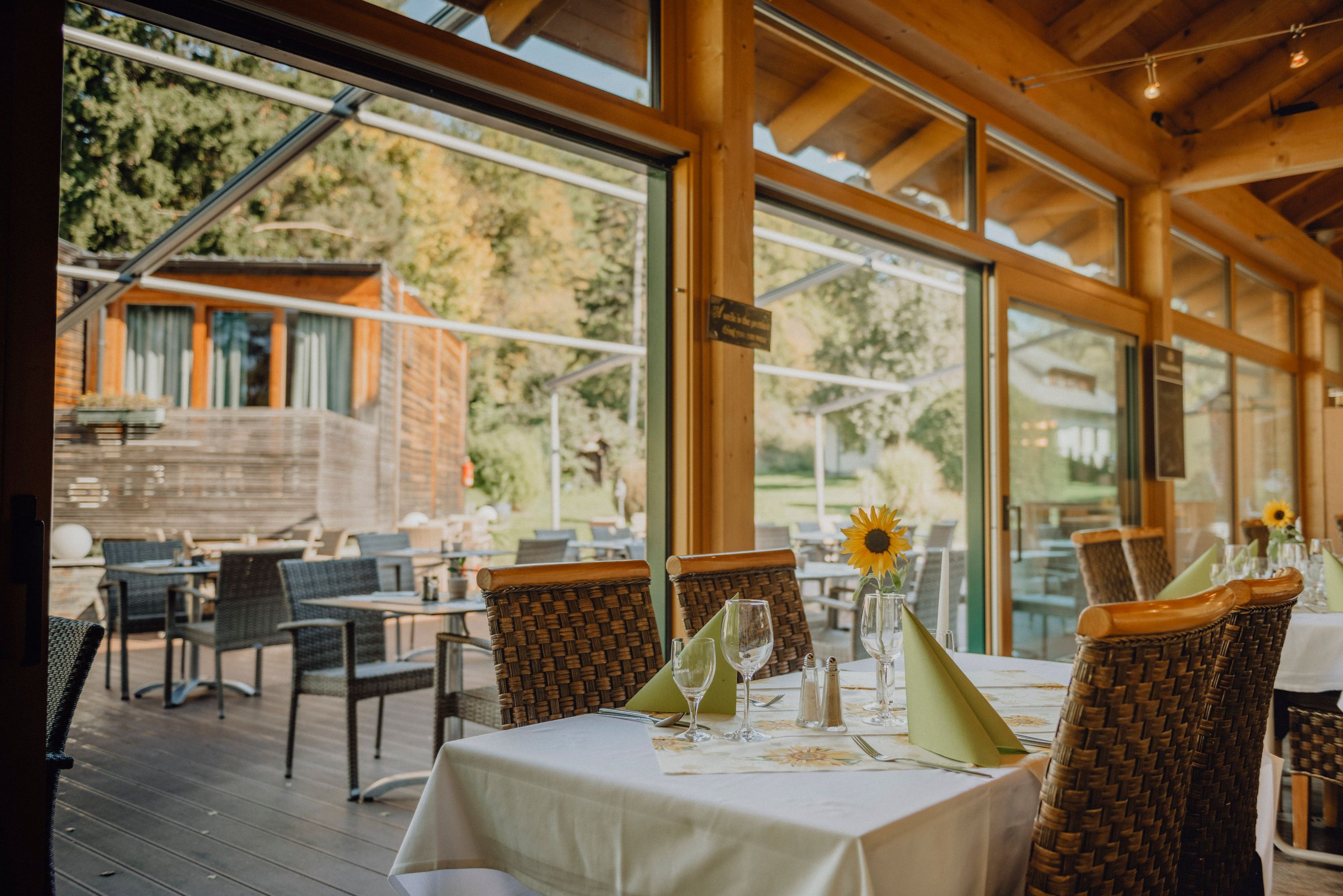 Ein Restaurant mit Wintergarten und Terrasse, gedeckte Tische mit Sonnenblumen, Blick auf grüne Umgebung.