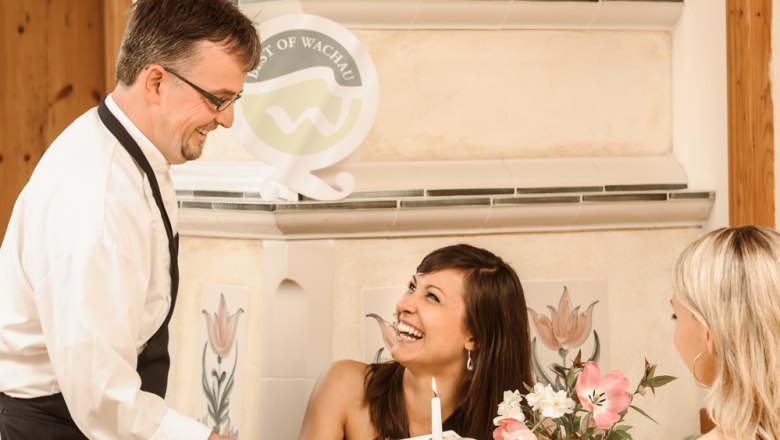 Ein Kellner serviert zwei Frauen in einem gemütlichen Restaurant. Eine Frau lacht, während sie das Essen entgegennimmt. Der Tisch ist elegant gedeckt.