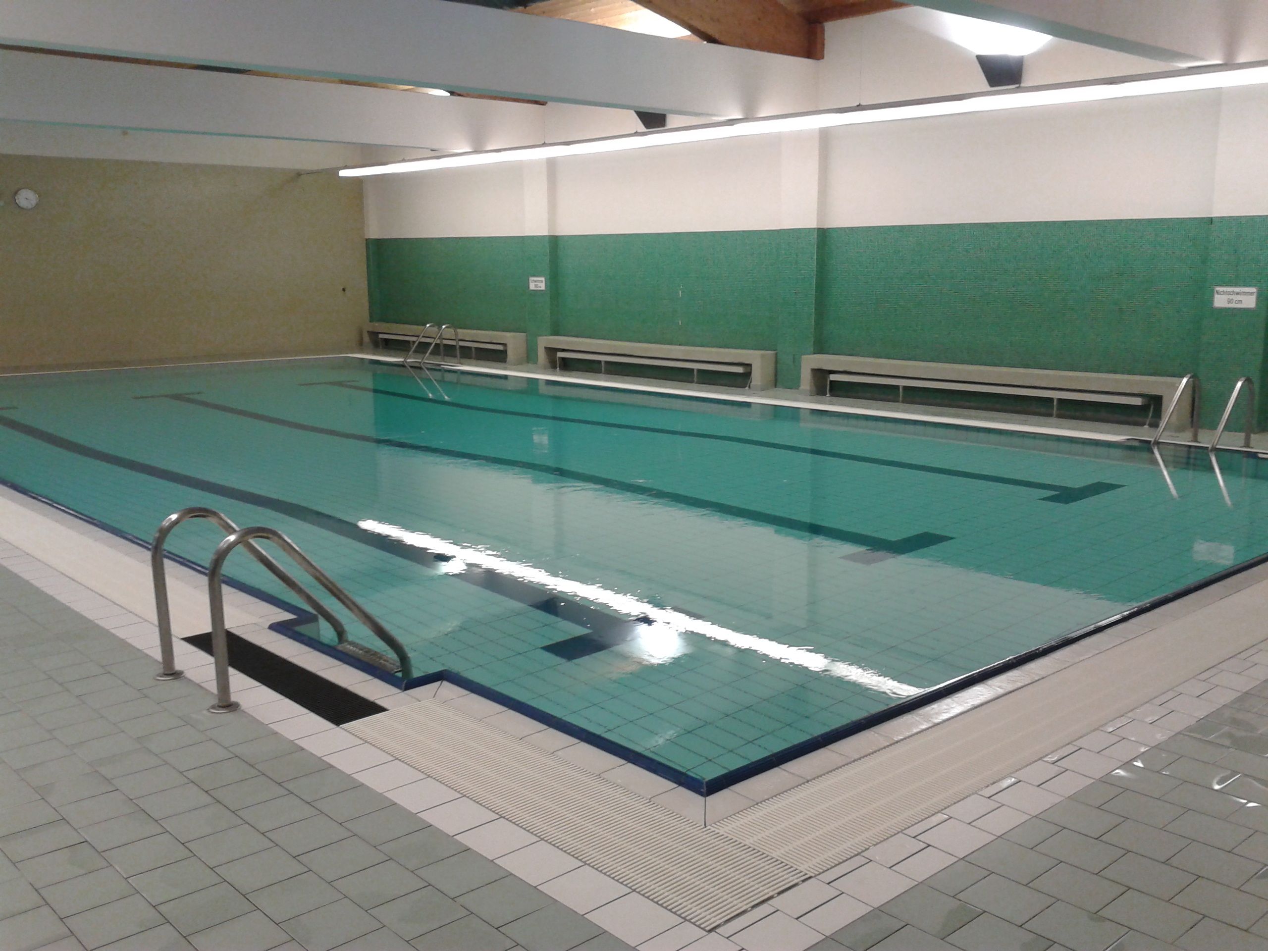 Innenansicht eines leeren Hallenbads mit grün gefliesten Wänden und einem großen Schwimmbecken.