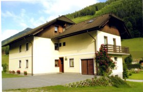 Ein zweistöckiges Haus mit Balkon und Rosenstrauch, umgeben von grünen Hügeln.