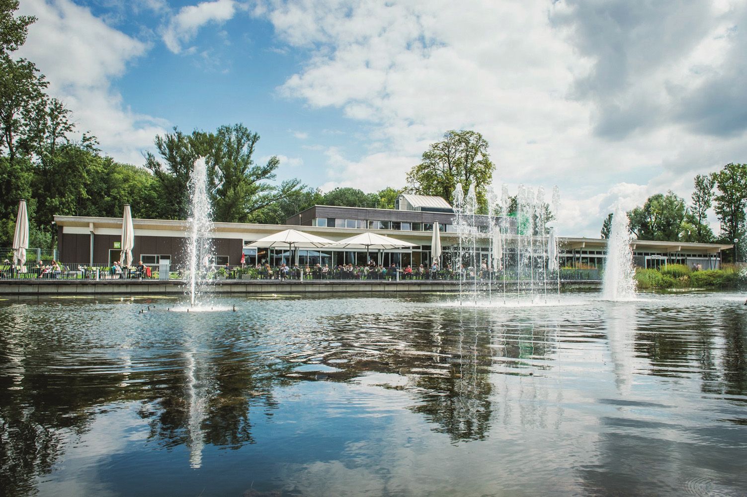 Ein modernes Gebäude mit Terrasse am Wasser, umgeben von Bäumen und Springbrunnen.