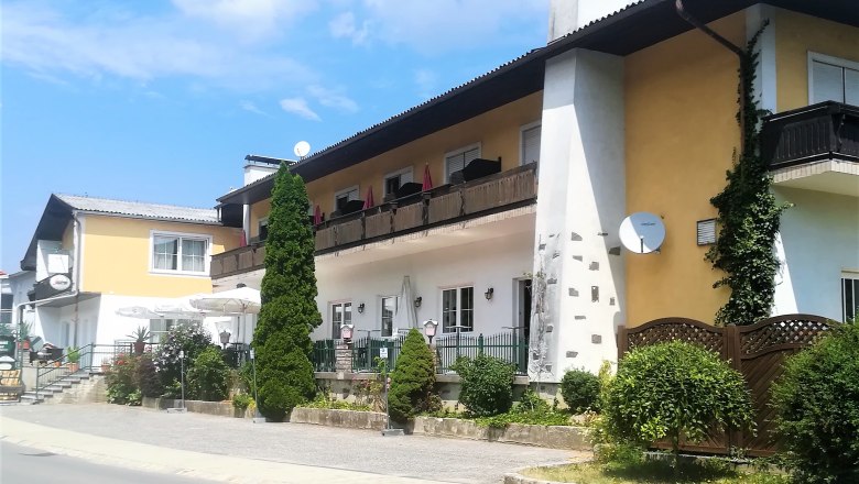 Ein zweistöckiges Gebäude mit Balkon und Terrasse, umgeben von Pflanzen und Bäumen, bei sonnigem Wetter.