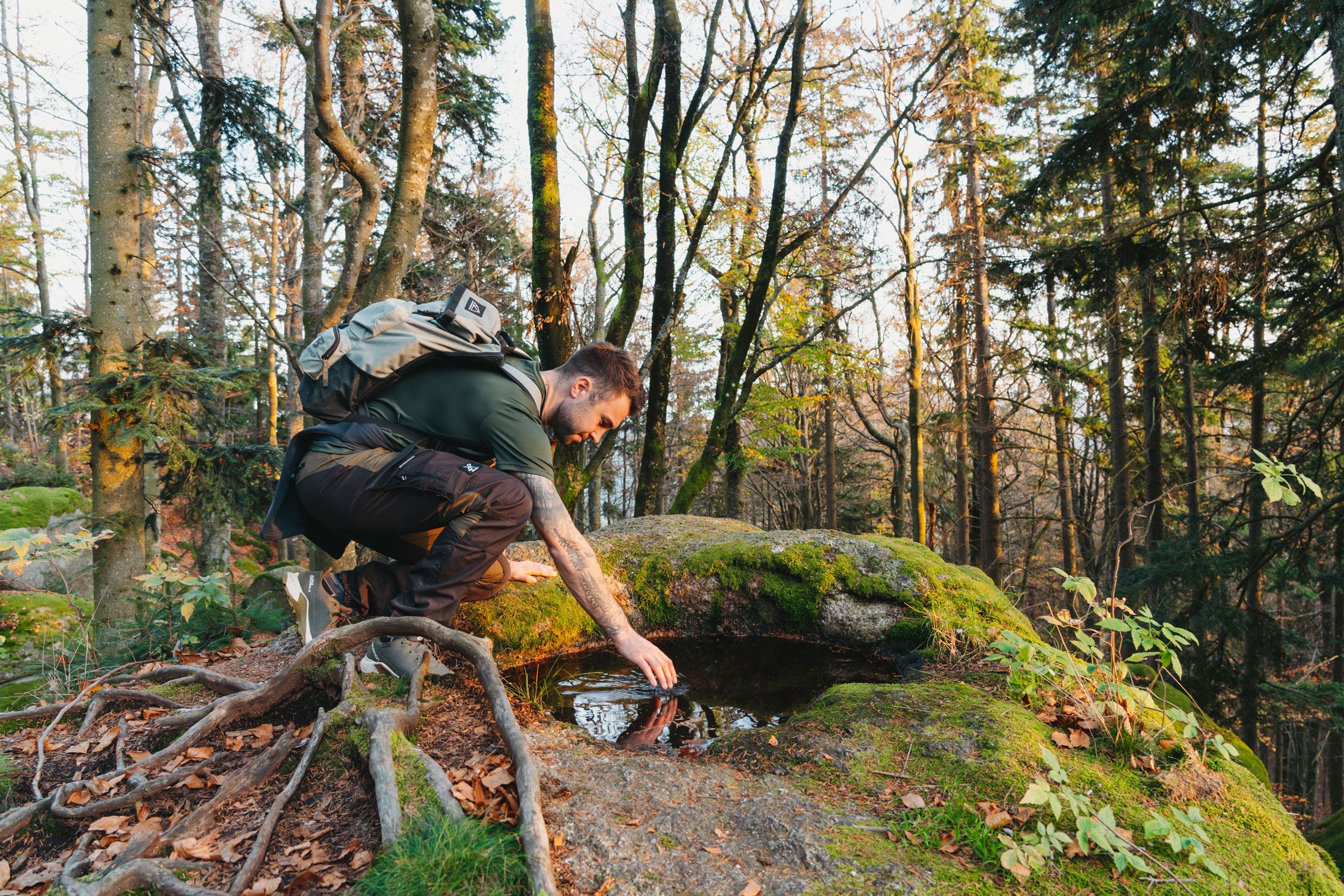 Ein Mann mit Rucksack kniet im Wald und berührt Wasser in einer Felsmulde.