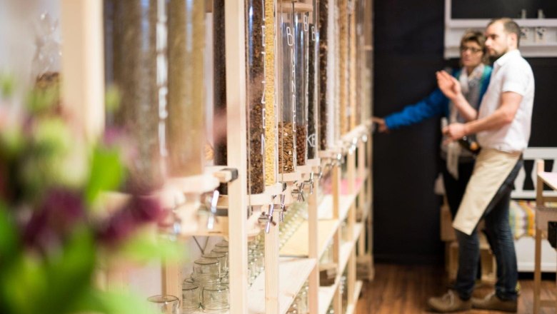 Innenansicht eines Unverpackt-Ladens mit Spendern f&uuml;r lose Lebensmittel und zwei Personen im Hintergrund.