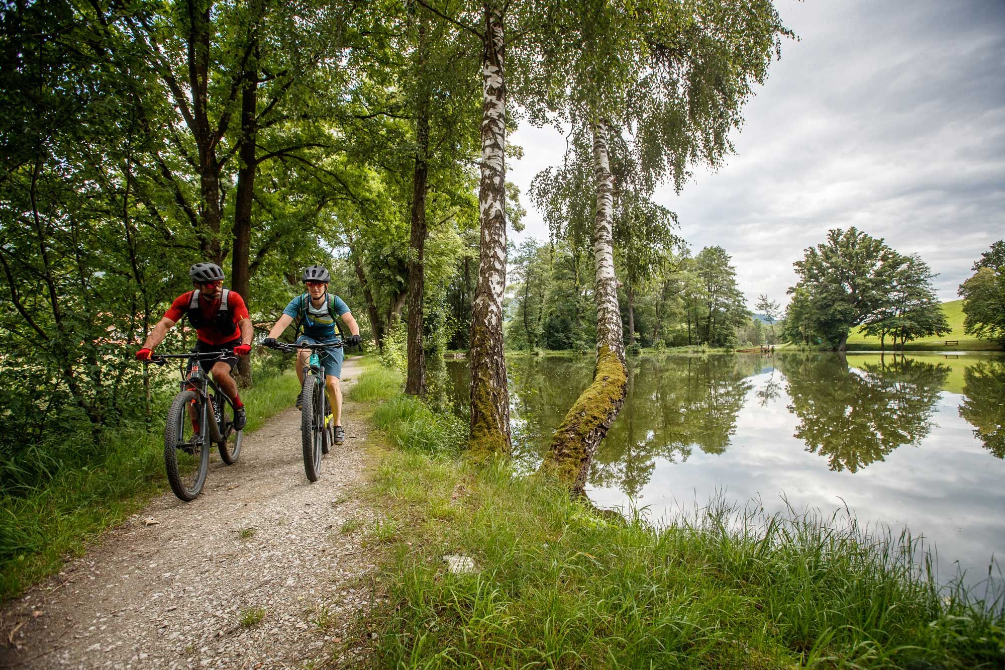 Zwei Mountainbiker fahren auf einem Waldweg neben einem ruhigen See.