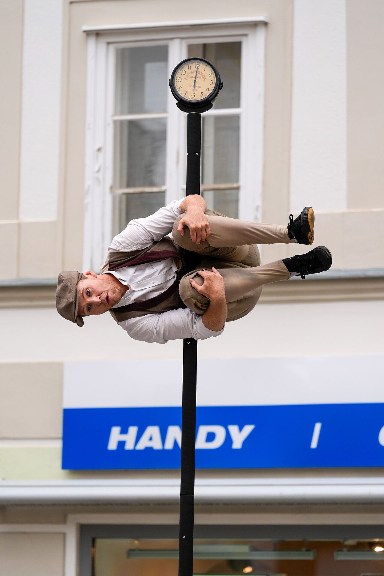 Straßenkünstler balanciert akrobatisch seitlich an einer hohen Stange mit Uhr.
