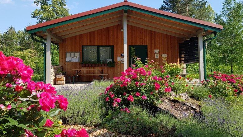 Holzgeb&auml;ude im Kr&auml;utergarten mit bl&uuml;henden Rosen und Lavendel im Vordergrund.