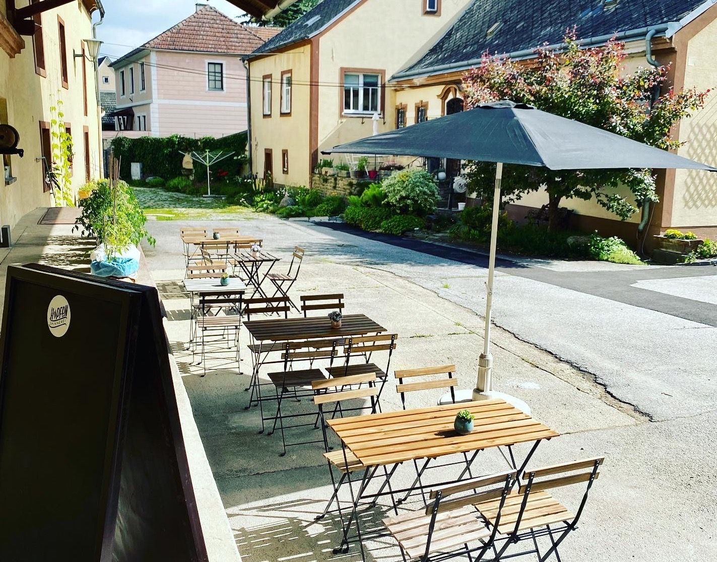 Ein Gastgarten mit Holztischen und Stühlen, einem Sonnenschirm und Pflanzen in einem Innenhof.