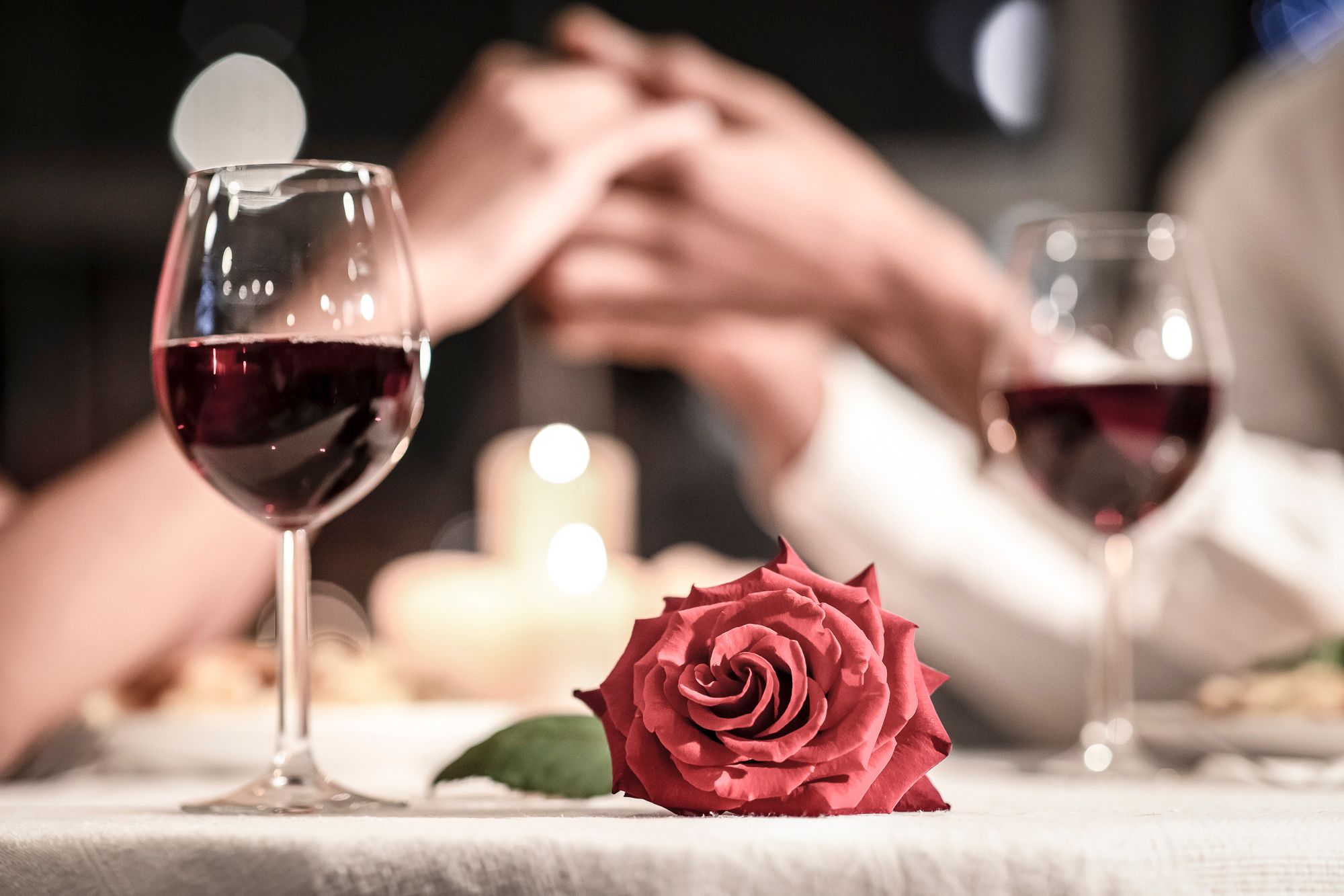 Zwei Weingläser, eine rote Rose und ein Paar, das sich die Hände hält, bei Kerzenschein.