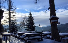 Verschneite Terrasse mit Holzb&auml;nken und Tischen, umgeben von B&auml;umen und Bergen im Hintergrund.