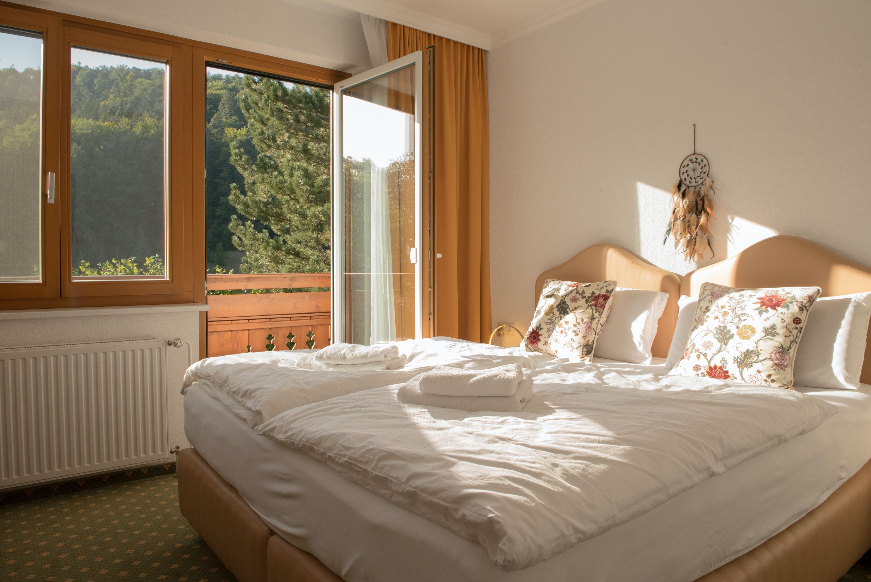 Ein gemütliches Einzelzimmer mit Doppelbett, dekorativen Kissen und einem offenen Balkon mit Blick auf grüne Bäume.