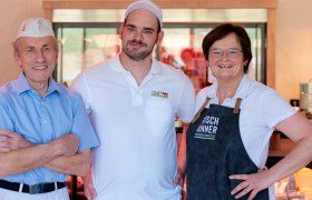 Drei l&auml;chelnde Personen in Arbeitskleidung stehen in einer Fleischerei.