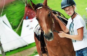 Ein Mädchen mit Helm steht neben einem braunen Pferd auf einem Bauernhof.