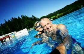 Ein &auml;lteres Paar schwimmt l&auml;chelnd in einem Freibad mit Wald im Hintergrund.