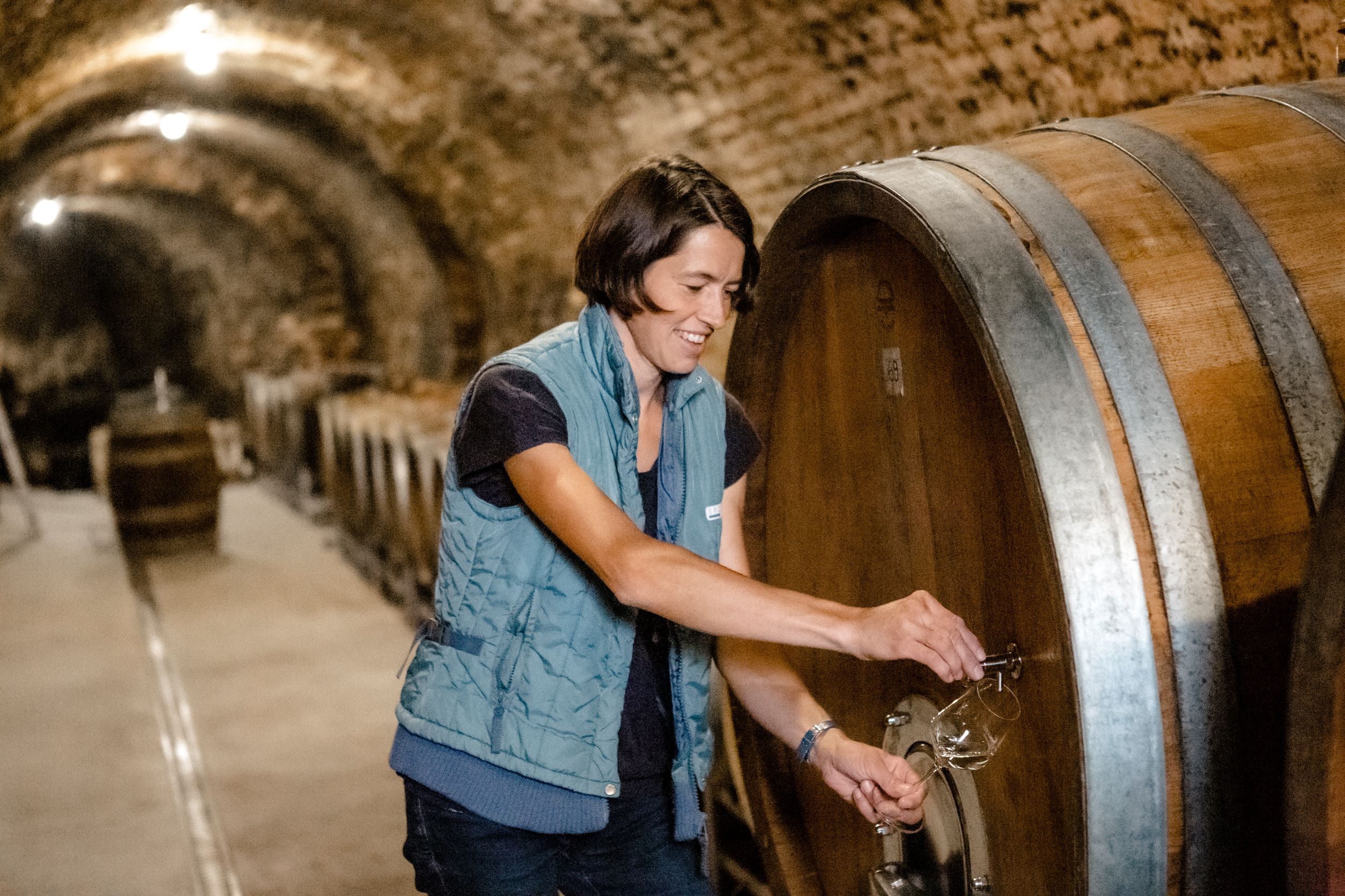 Person in einer Weinkellerei zapft Wein aus einem großen Holzfass.
