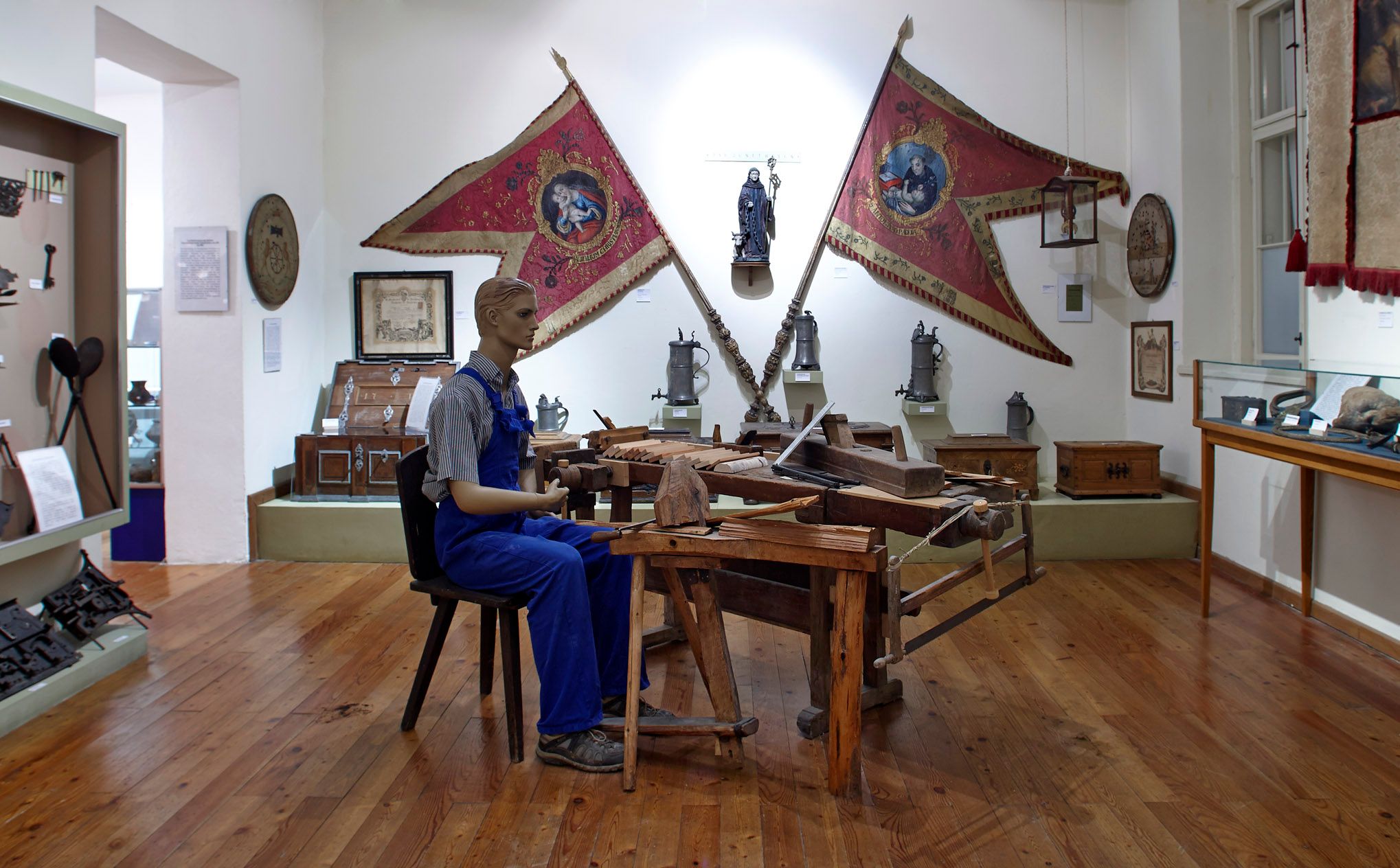 Innenansicht eines Museums mit einer Schaufensterpuppe in Arbeitskleidung an einem Holztisch, umgeben von historischen Gegenständen und Fahnen.