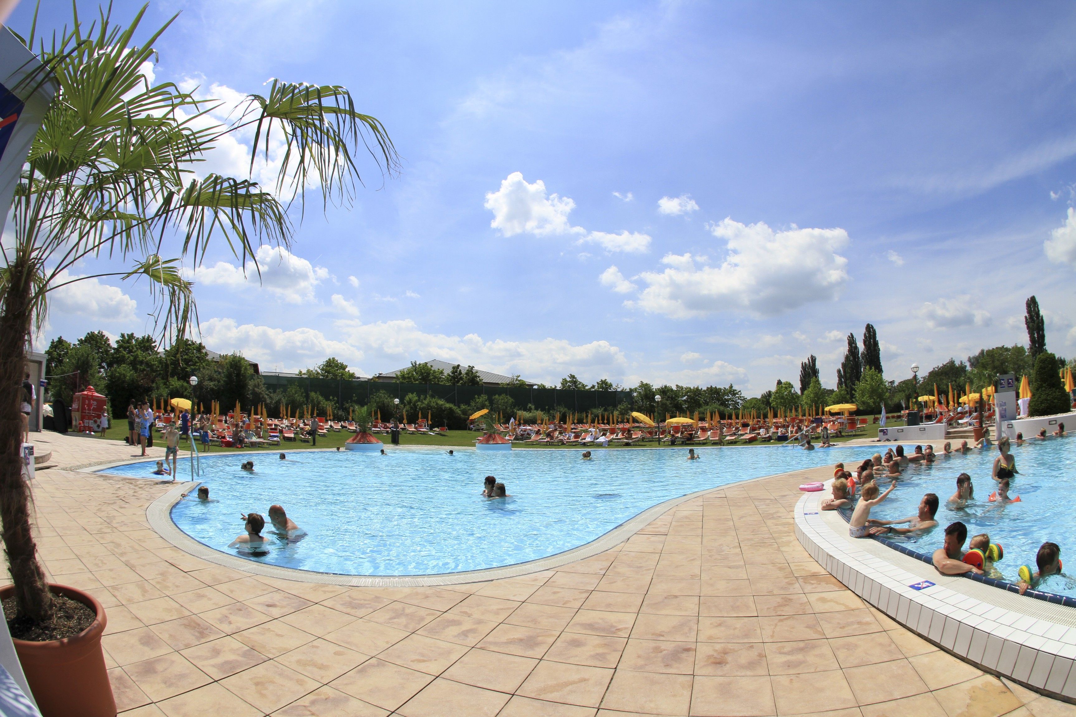 Ein Außenpool in einem Sommerbad mit vielen Menschen, umgeben von Liegestühlen und Sonnenschirmen unter einem blauen Himmel.