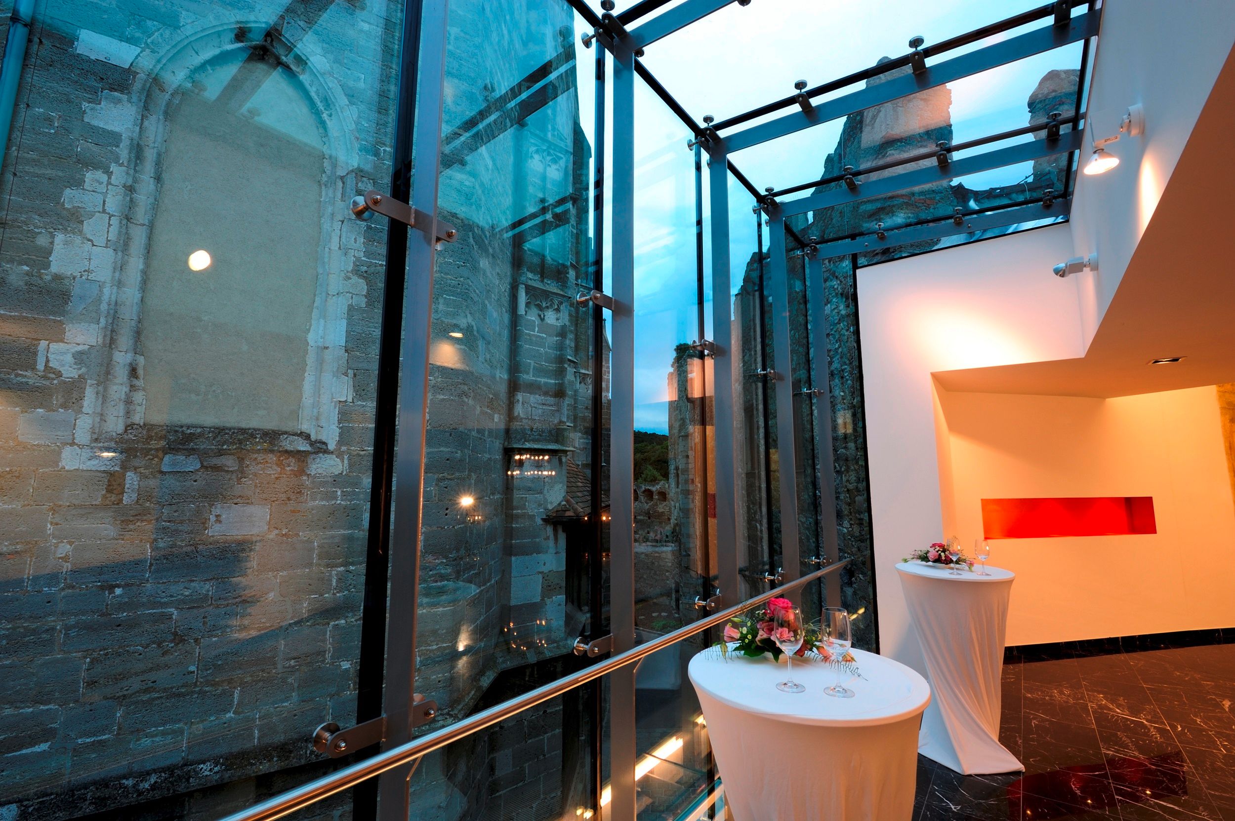 Moderne Glasarchitektur neben alter Steinmauer mit dekorierten Stehtischen.