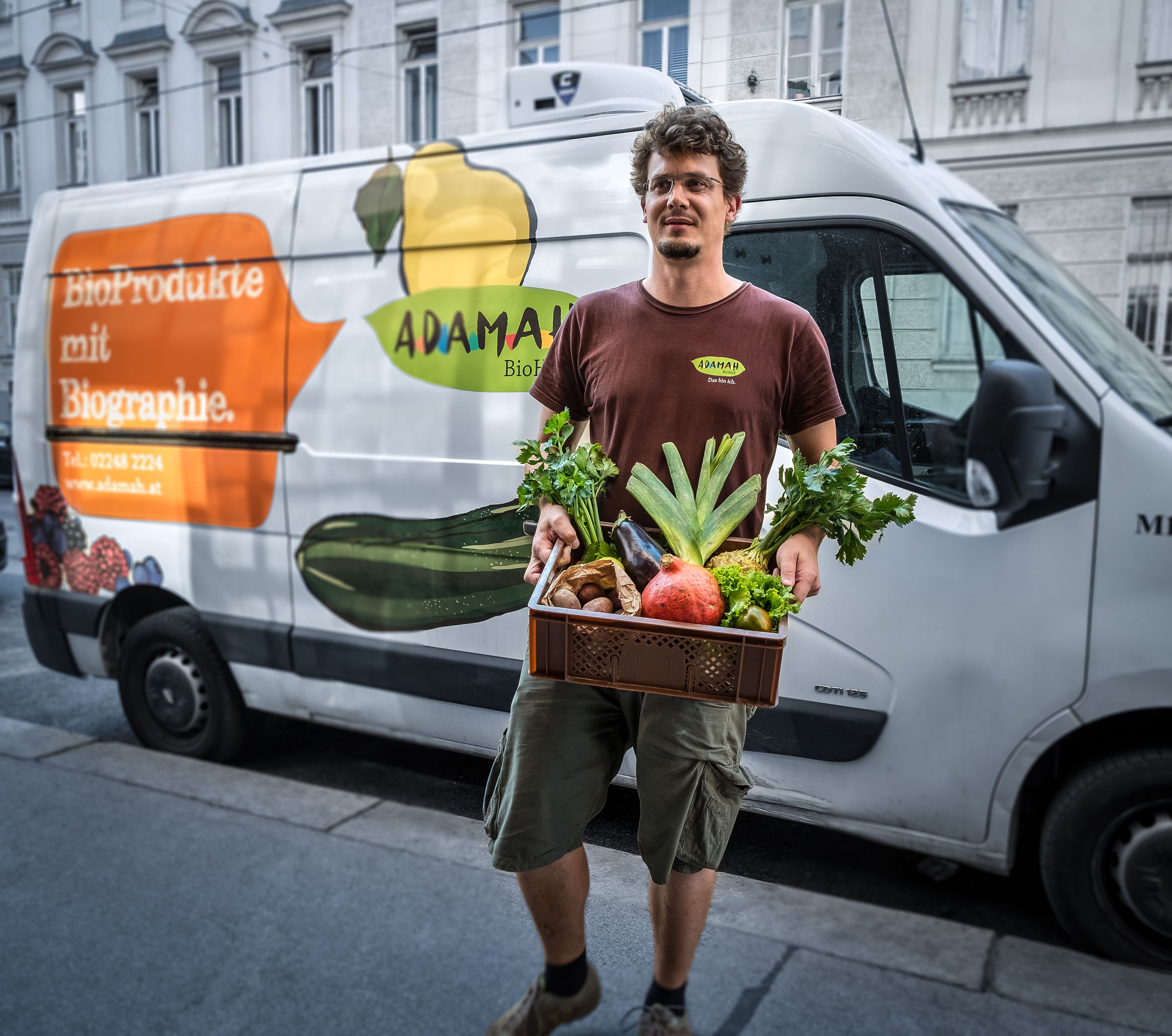 Ein Mann hält eine Kiste mit frischem Obst und Gemüse vor einem Lieferwagen mit ADAMAH-Logo.