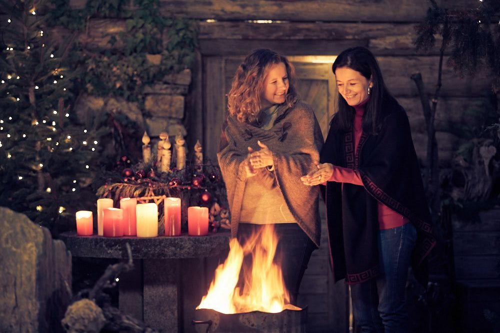 Zwei Frauen stehen lächelnd an einem Feuer, umgeben von Kerzen und Weihnachtsdekoration.