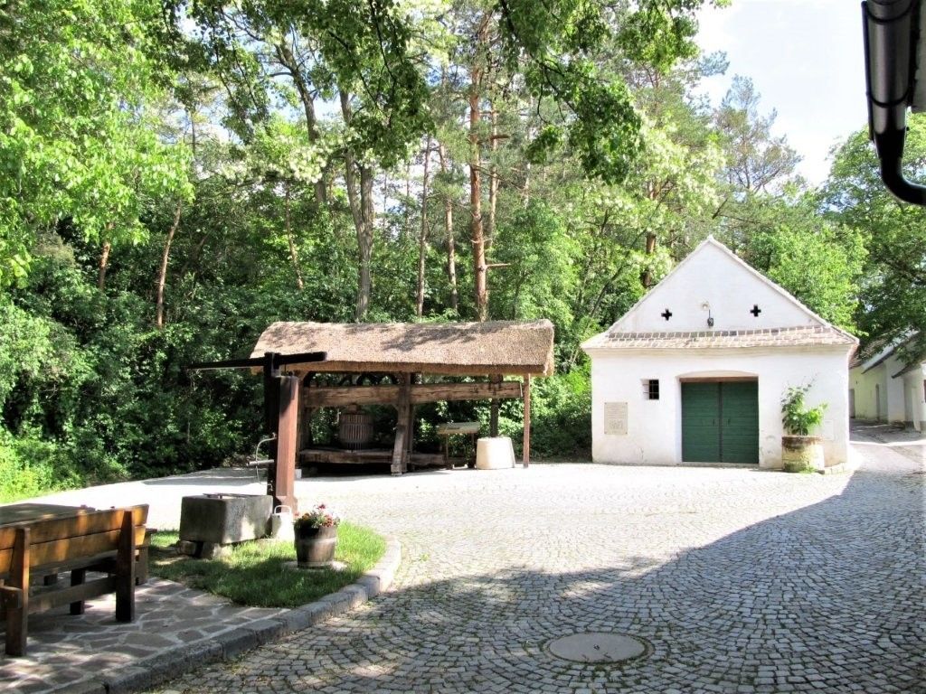 Ein gepflasterter Hof mit einem kleinen weißen Gebäude und einem überdachten Holzgestell, umgeben von Bäumen.