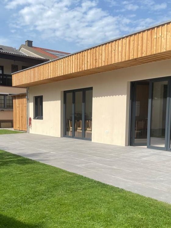 Moderne Gebäudeaußenansicht mit Holzverkleidung und großen Fenstern, umgeben von einer Terrasse und Rasenfläche.