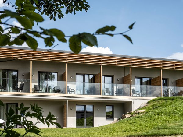 Modernes zweistöckiges Gebäude mit Glasbalkonen und Holzverkleidung, umgeben von grüner Landschaft.