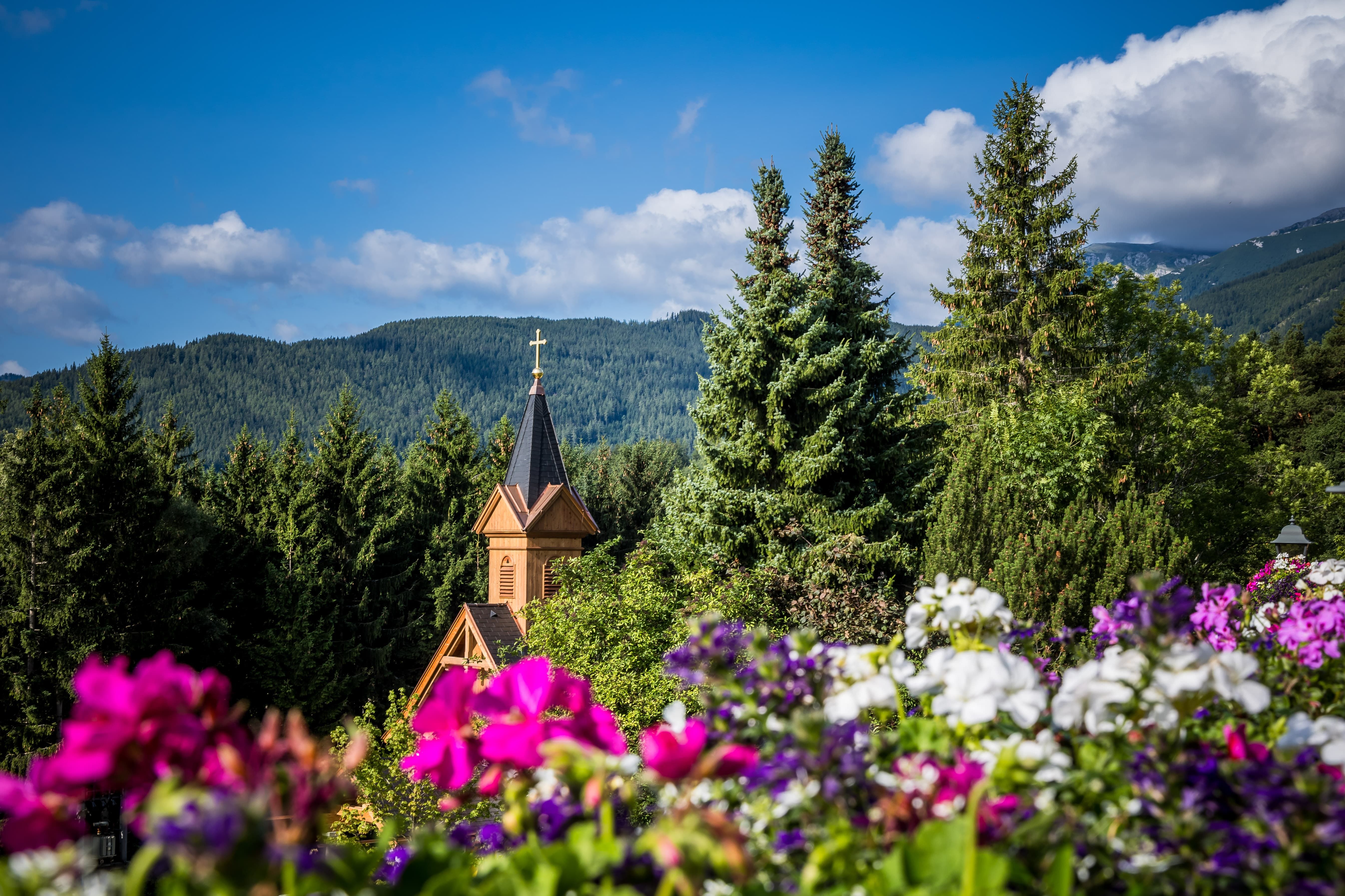 Kirchturm inmitten von Bäumen und Blumen vor einer Berglandschaft.