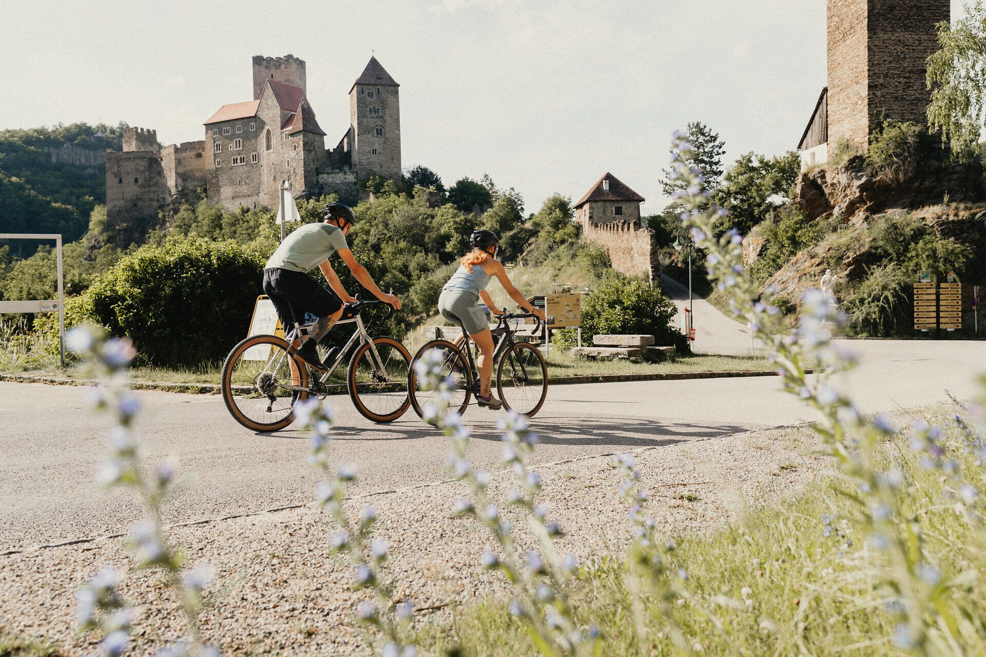 Zwei Radfahrer fahren entlang eine Straße, hinter ihnen die Burg Hardegg, im Vordergrund Blumen