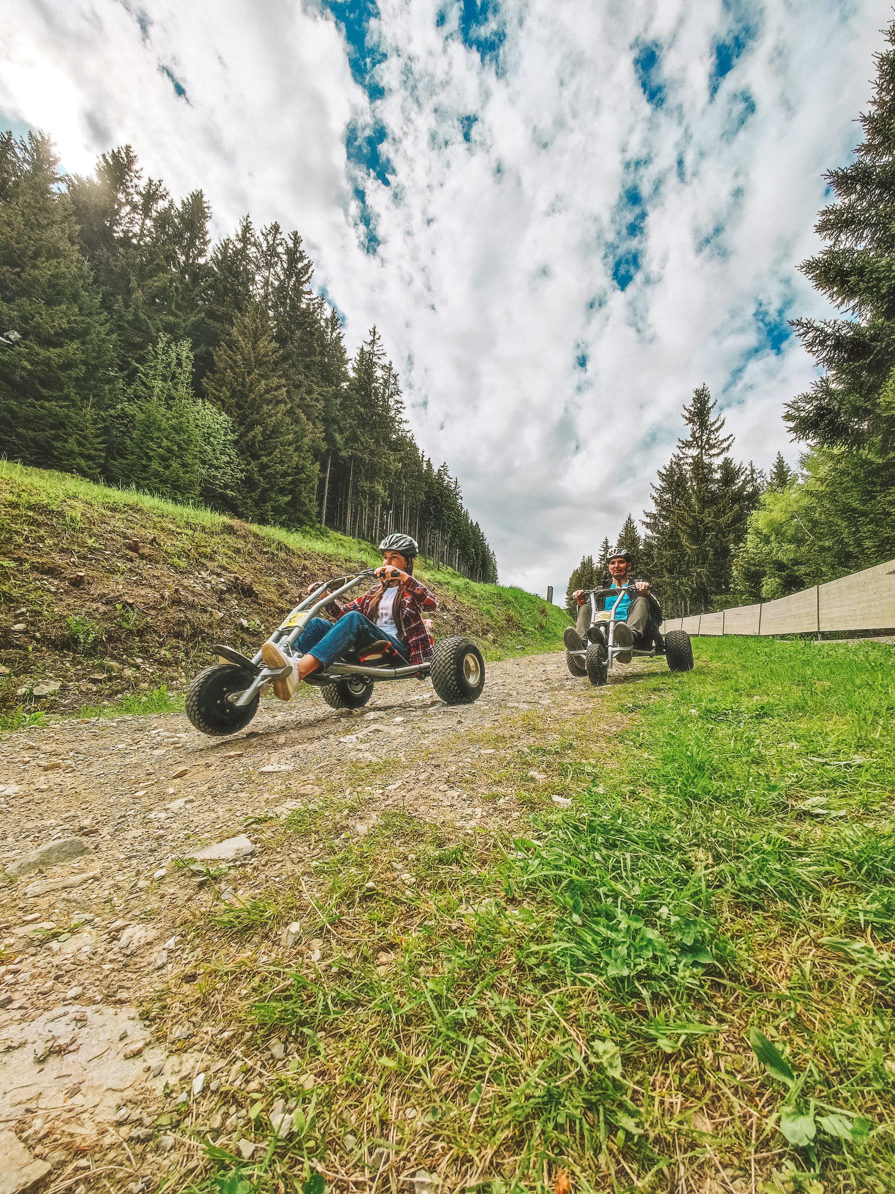 Zwei Personen fahren mit Mountaincarts auf einem Schotterweg im Wald.