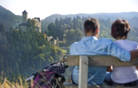Ein Paar sitzt auf einer Bank mit Blick auf eine Burg in einer bewaldeten Landschaft.