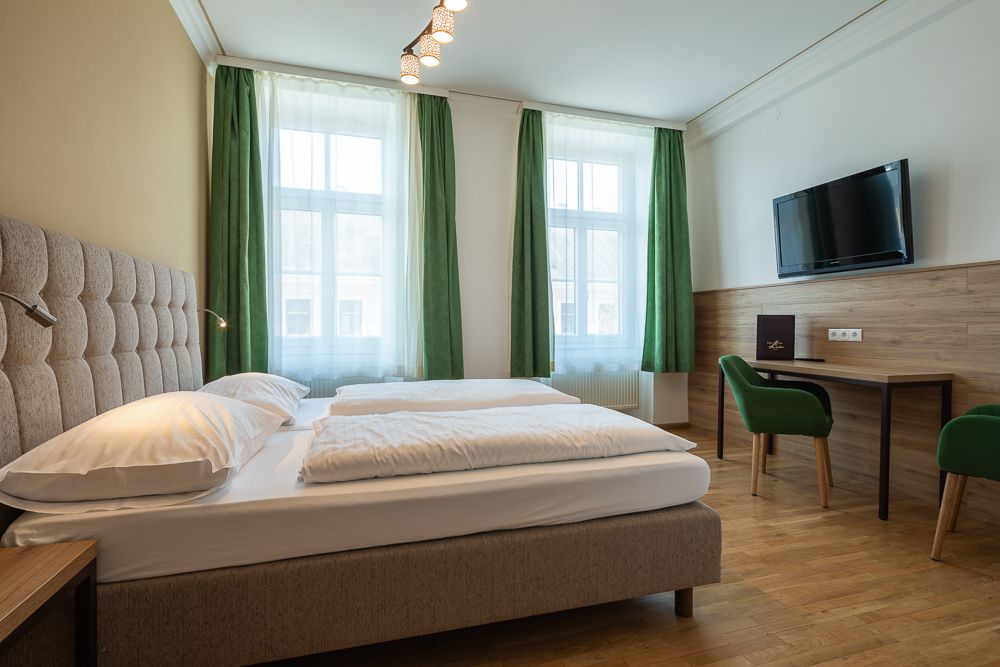 Hotelzimmer mit Doppelbett, grüne Vorhänge, Schreibtisch, Stuhl und Wand-TV.