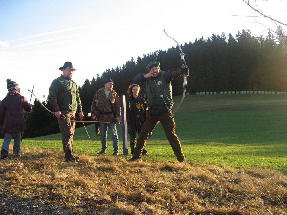 Gruppe von Menschen beim Bogenschießen auf einer Wiese vor einem Wald.