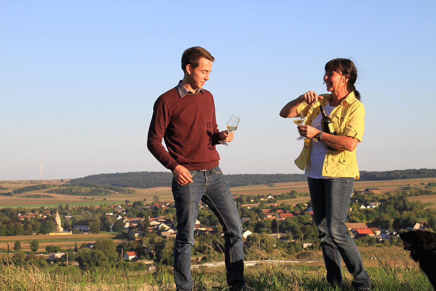 Zwei Personen stehen auf einem Hügel mit Weingläsern in der Hand, im Hintergrund eine ländliche Landschaft mit Windrädern.