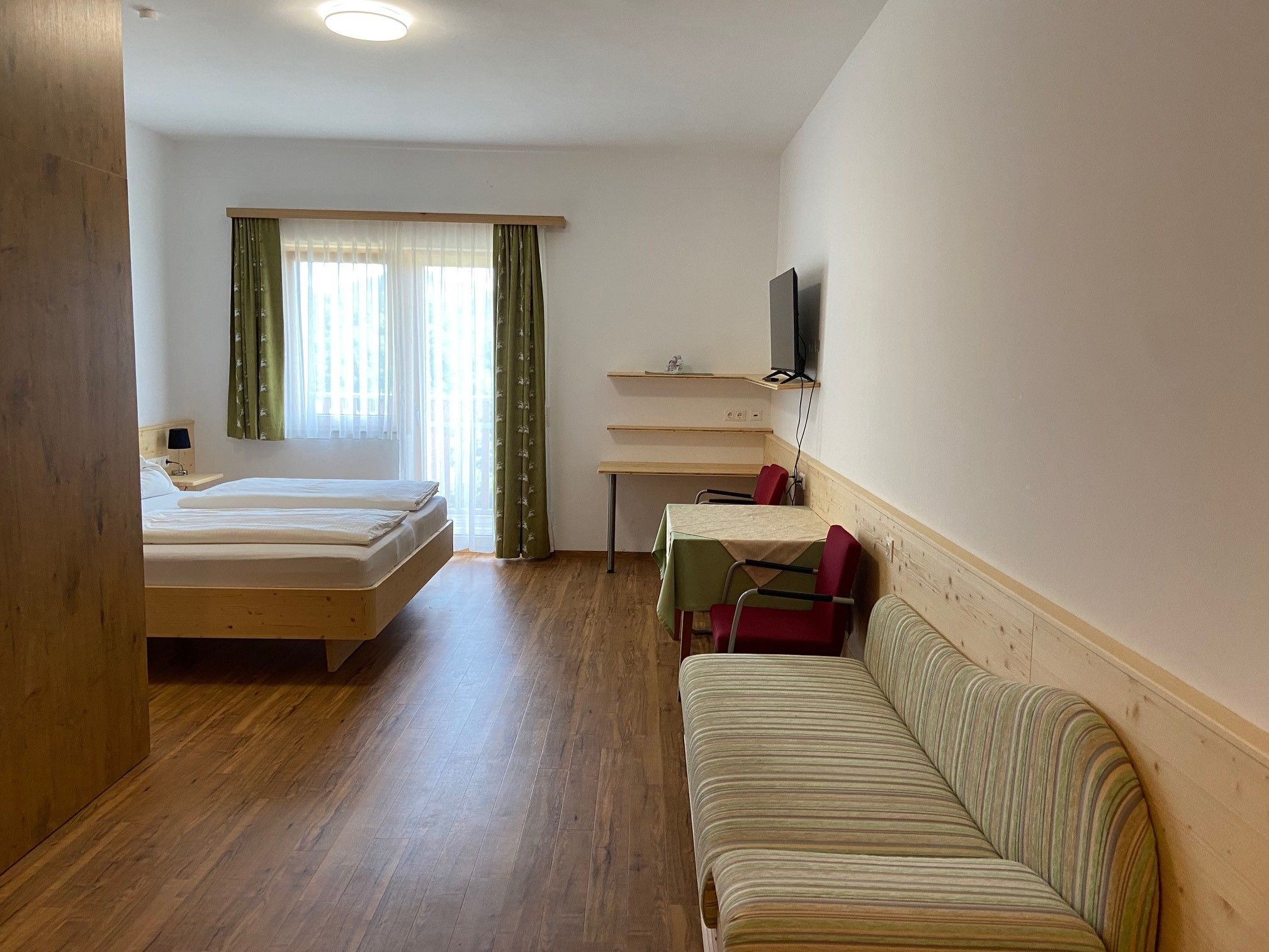 Ein modernes Hotelzimmer mit Doppelbett, Sofa, Tisch und Stühlen. Ein Fernseher hängt an der Wand. Vorhänge an einem Fenster lassen Tageslicht herein.