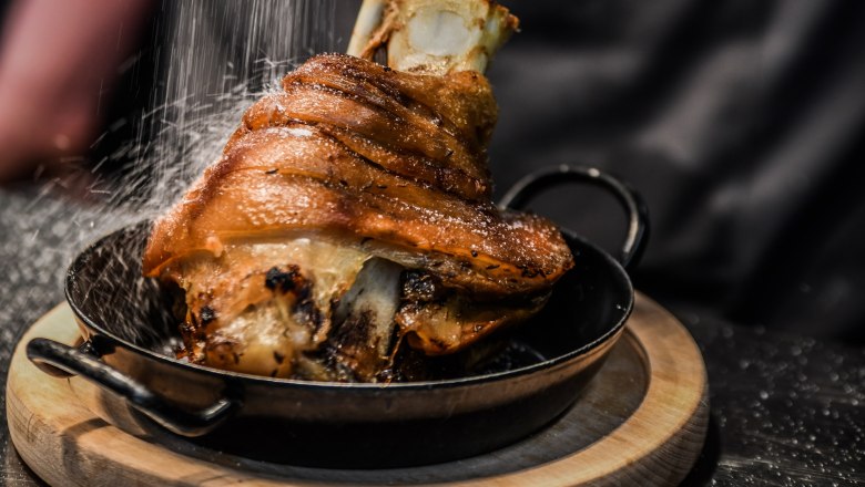 Knusprige Schweinshaxe in einer Pfanne auf einem Holzbrett, mit Salz bestreut.