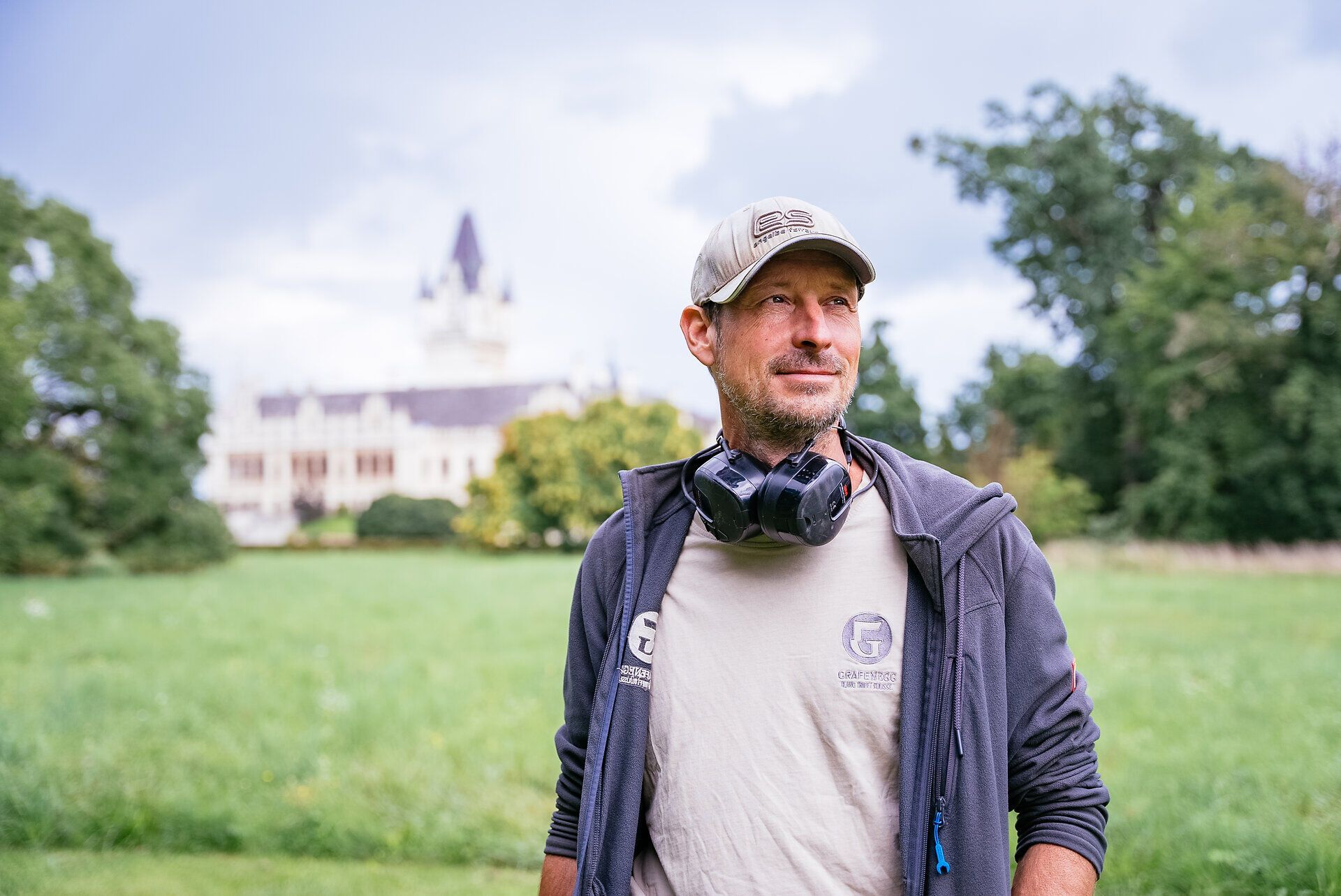 Alexander Malik steht in lockerem Outfit mit Händen in den Hosentaschen in einer Wiese vor Schloss Grafenegg