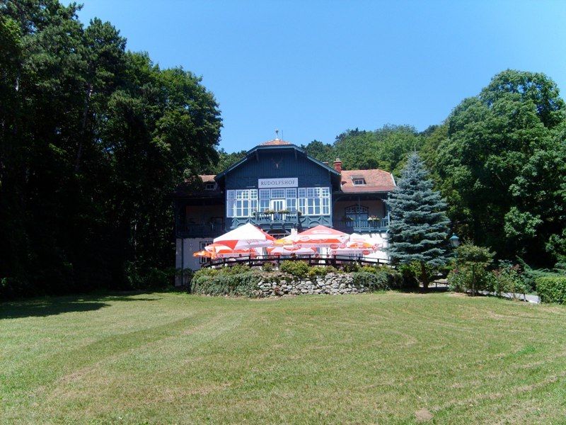 Ein großes Haus namens Rudolfshof mit Terrasse und Sonnenschirmen, umgeben von Bäumen und einer Wiese.