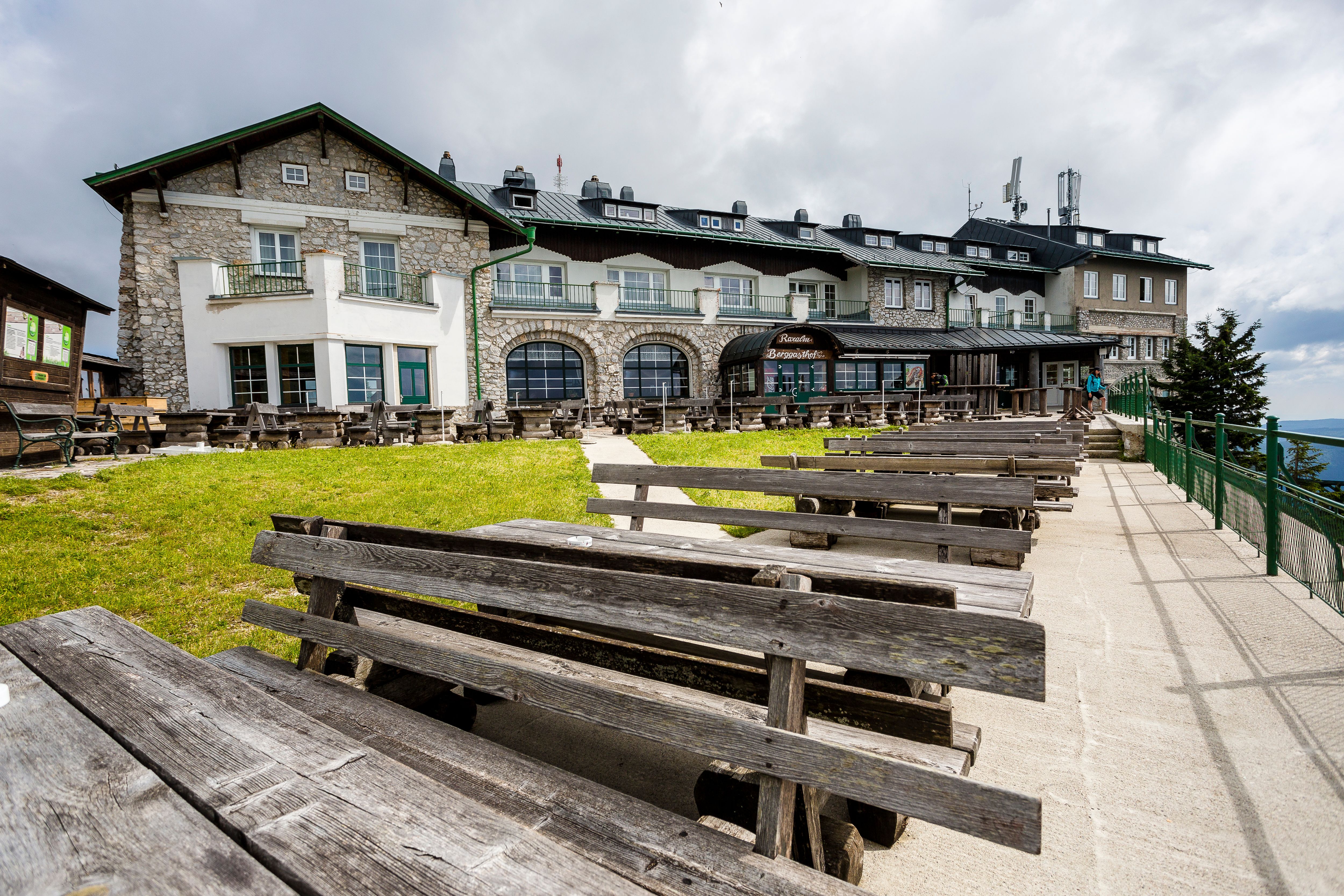 Ein Berggasthof mit Holzbänken und Tischen im Freien, umgeben von grüner Wiese und bewölktem Himmel.
