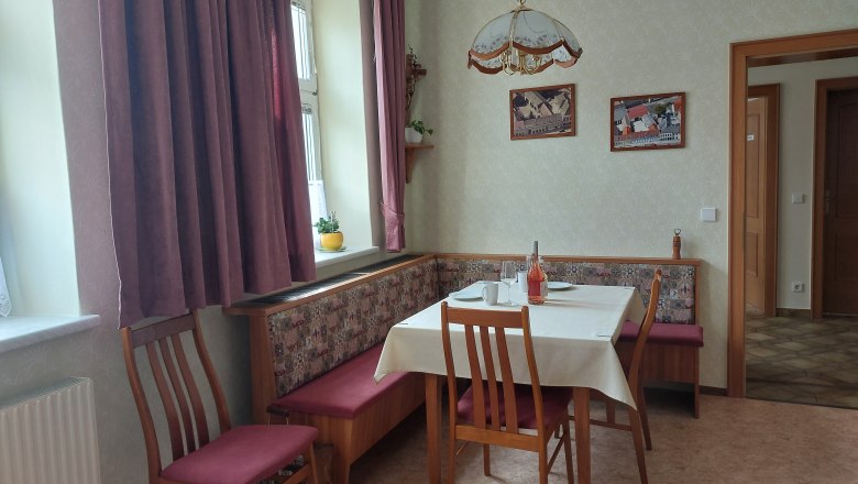 Gemütliche Wohnküche mit Esstisch, Stühlen und Eckbank. Fenster mit Vorhängen, Bilder an der Wand.