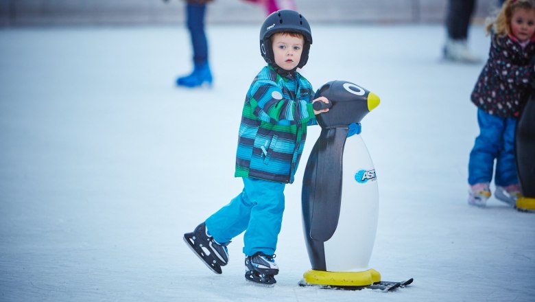 Ein Kind mit Helm und bunter Jacke h&auml;lt sich an einer Pinguin-Eislaufhilfe auf einer Eisbahn fest.