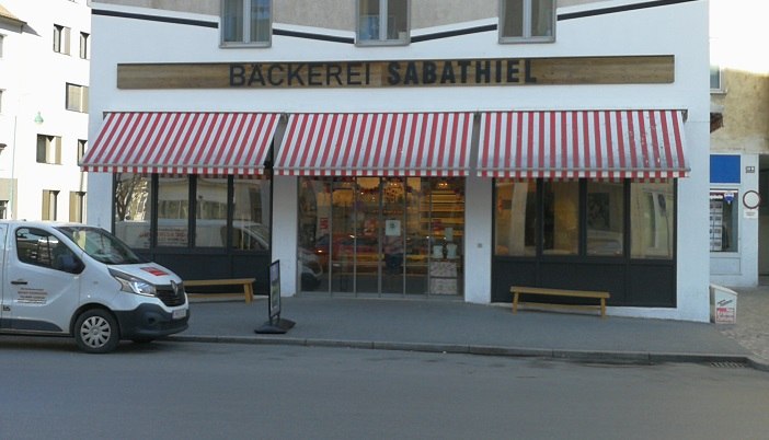 Au&szlig;enansicht der B&auml;ckerei Sabathiel mit rot-wei&szlig; gestreifter Markise und Lieferwagen davor.