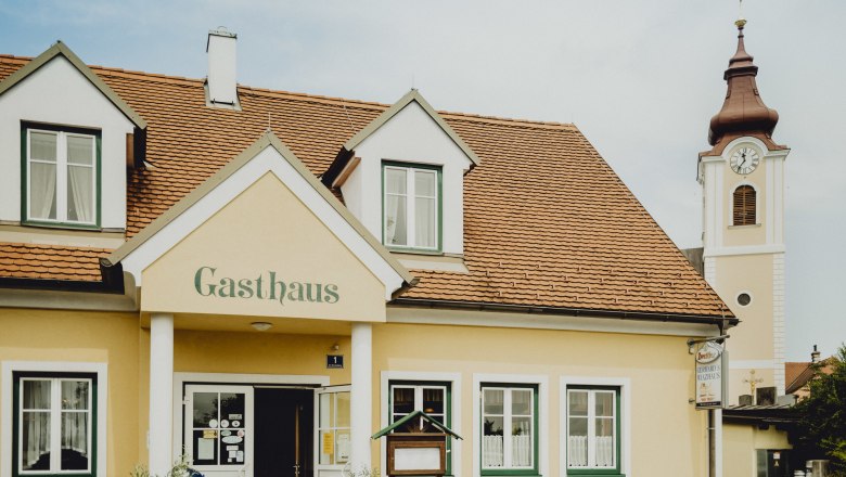Ein gelbes Gasthaus mit roten Dachziegeln und einem Kirchturm im Hintergrund.