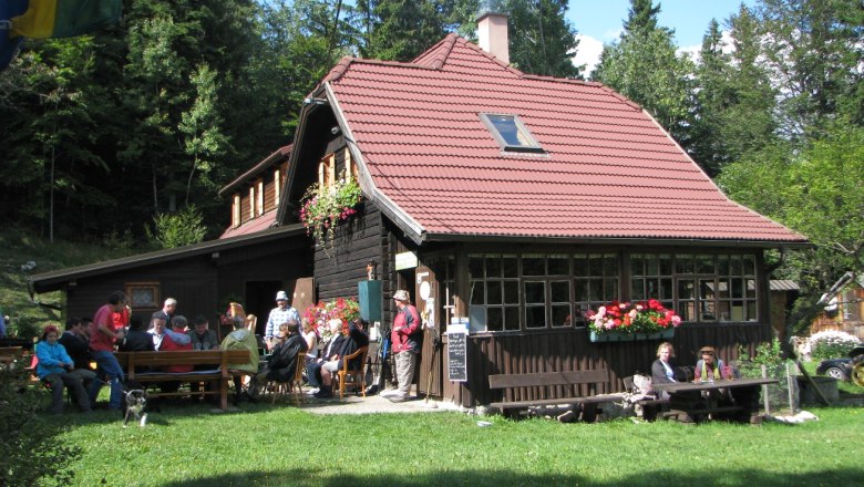 Eine Berghütte mit rotem Dach, umgeben von Wald, mit Menschen, die draußen sitzen.