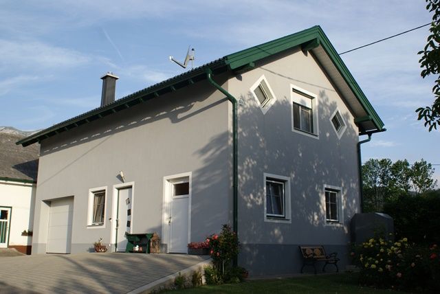 Graues zweistöckiges Haus mit grünem Dach und Gartenbank.