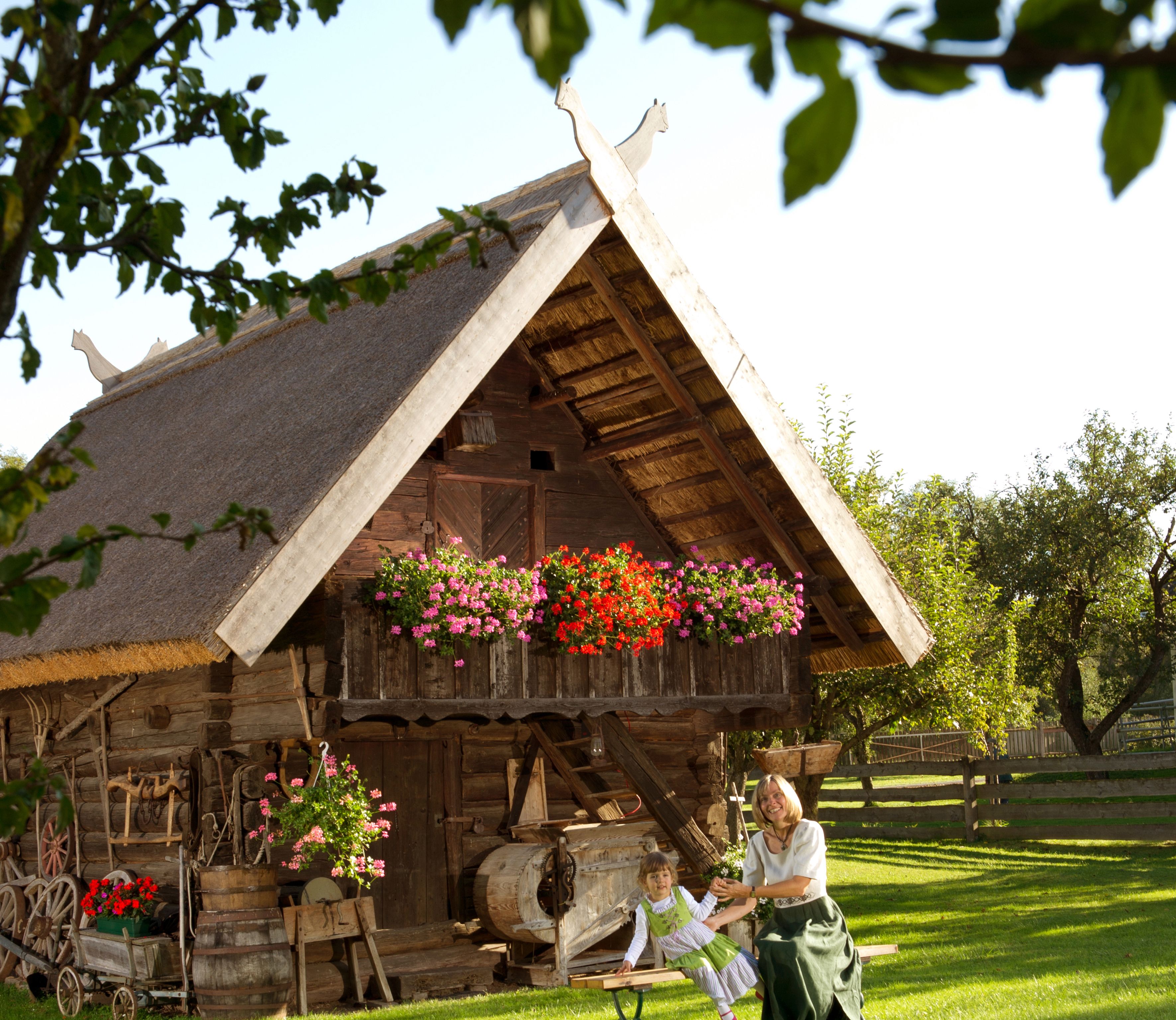 Traditionelles Holzgebäude mit Strohdach und Blumen, Frau und Kind in Tracht davor.