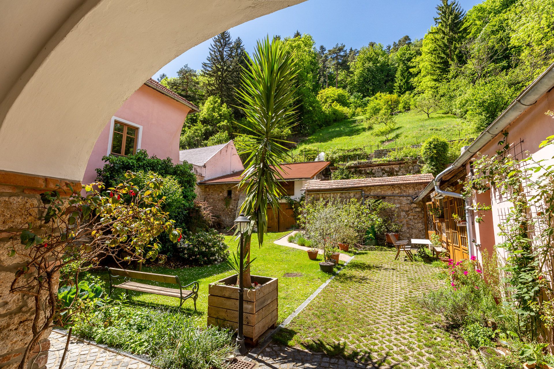 Ein idyllischer Innenhof mit Pflanzen, Bänken und einem Tisch, umgeben von rosa Gebäuden und grünen Hügeln.